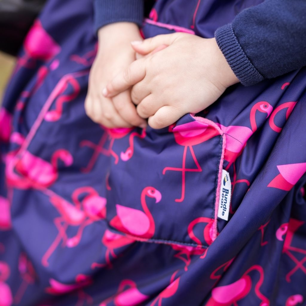Kids fleece-lined wheelchair cosies. Waterproof, warm and easy to fit. Seen here in a flamingo design. and showing the hand warming pocket.